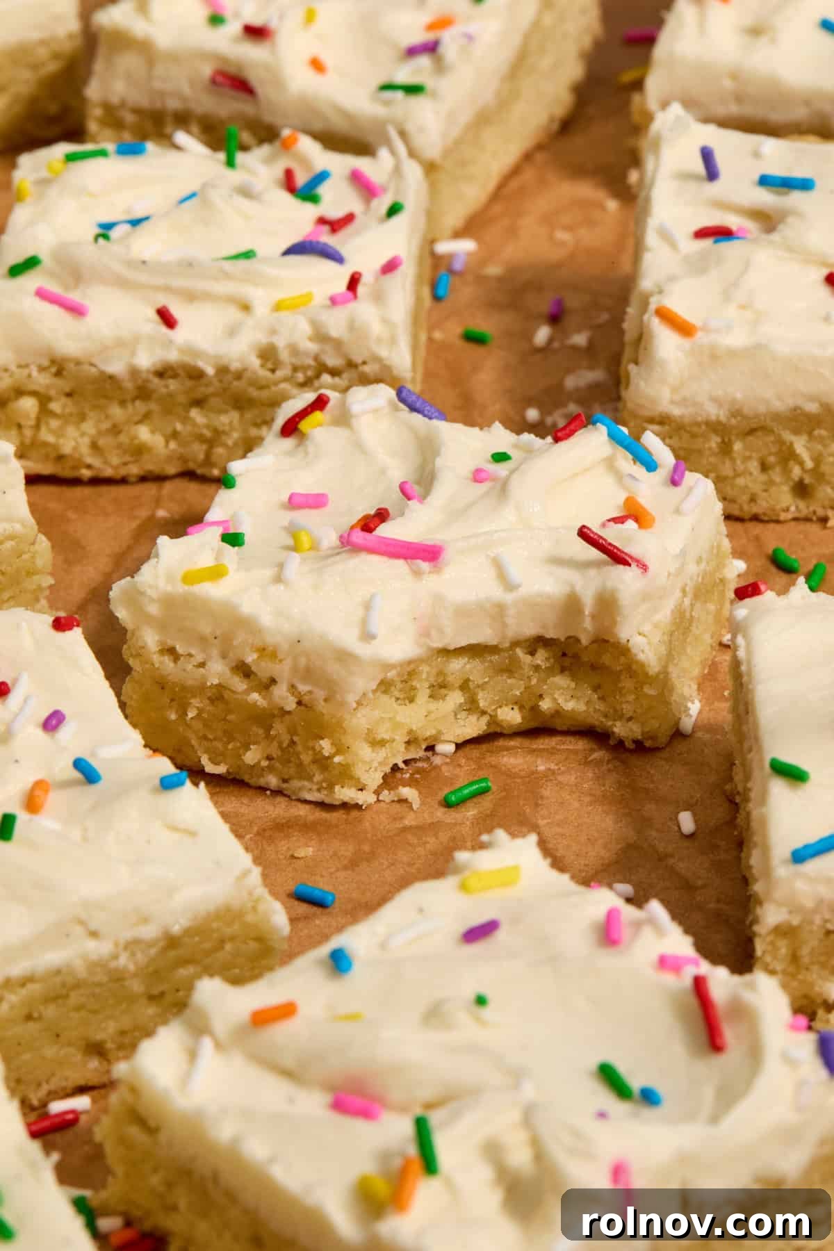 Decadent vanilla frosted sugar cookie bars adorned with vibrant rainbow sprinkles, elegantly arranged on parchment paper.
