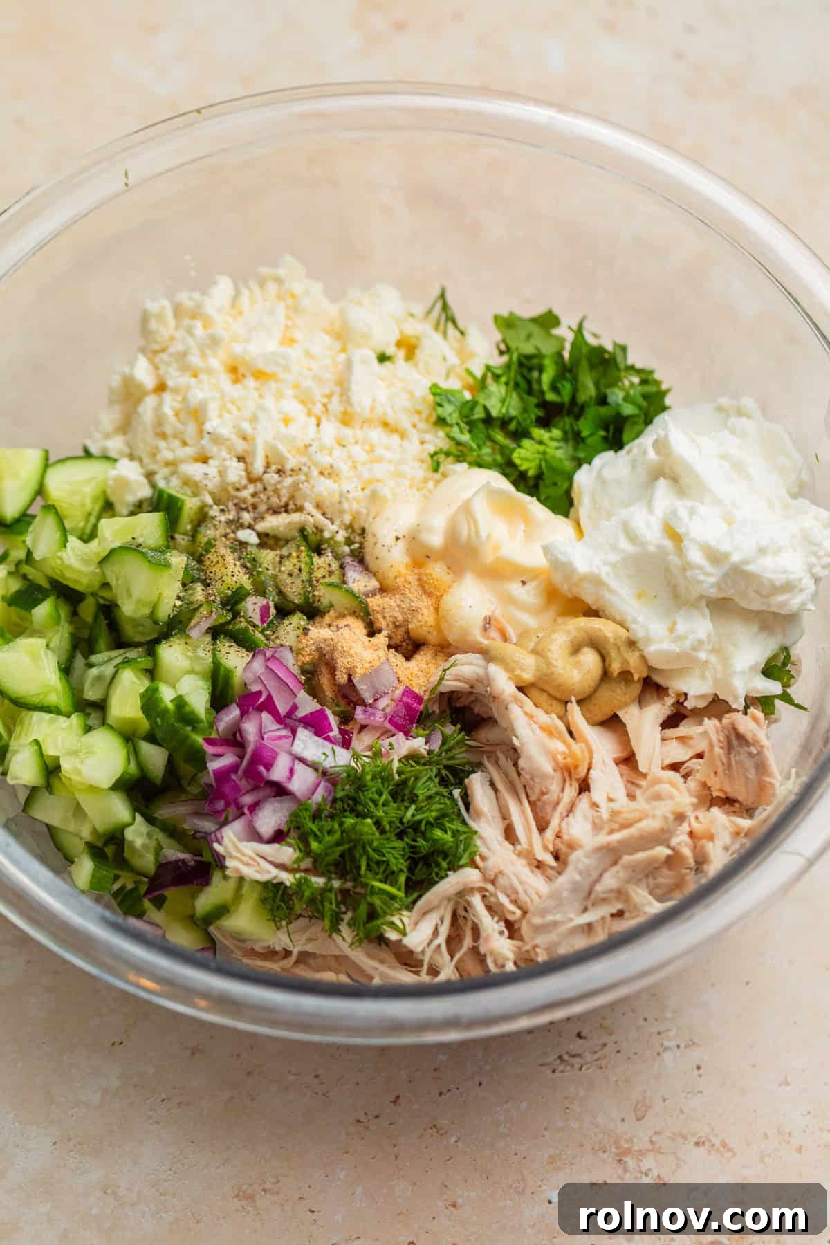 Shredded chicken, finely diced cucumbers, crumbled feta cheese, creamy Greek yogurt, and other seasonings and herbs are combined in a large mixing bowl, ready to be mixed.