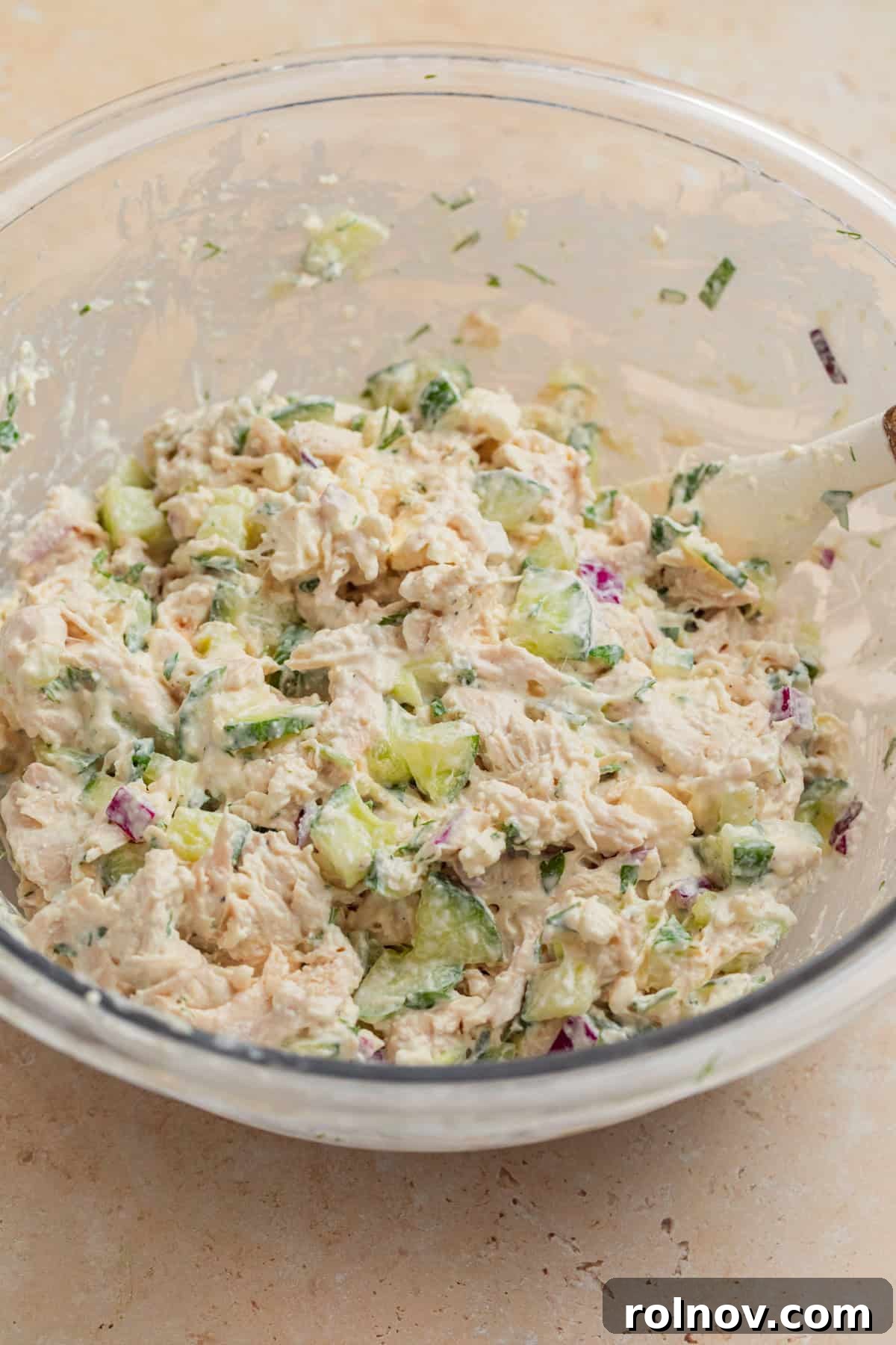 The mixed cucumber and feta chicken salad in a bowl, with a spatula used to combine all ingredients, showing its creamy and chunky texture.