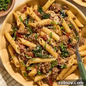 A plate of vibrant sun-dried tomato pasta with Italian sausage, garnished with fresh Parmesan and chopped basil.