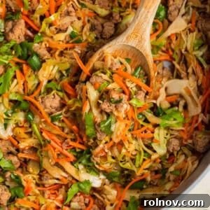 A large pot filled with a Ground Turkey Egg Roll Bowl, featuring a medley of ingredients and a wooden spoon resting inside.