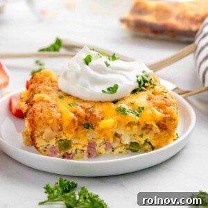 A slice of hearty breakfast casserole with crispy tater tots, garnished with fresh parsley and a dollop of sour cream on a white plate.