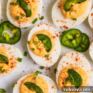 A festive serving dish filled with spicy deviled eggs, garnished with finely chopped chives, fresh jalapeño slices, and a dusting of paprika.
