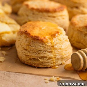 Golden-brown honey biscuits resting on tan parchment paper, with luscious honey drizzling over the top from a honey dipper stick positioned beside them.