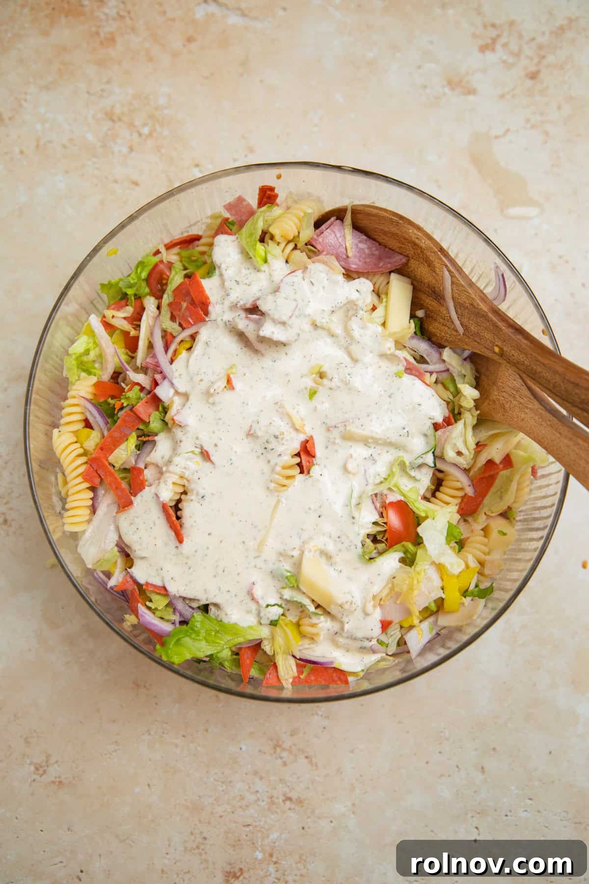 Loaded Grinder Pasta Salad 9 Creamy parmesan dressing being poured generously over the Grinder Pasta Salad in a large bowl.