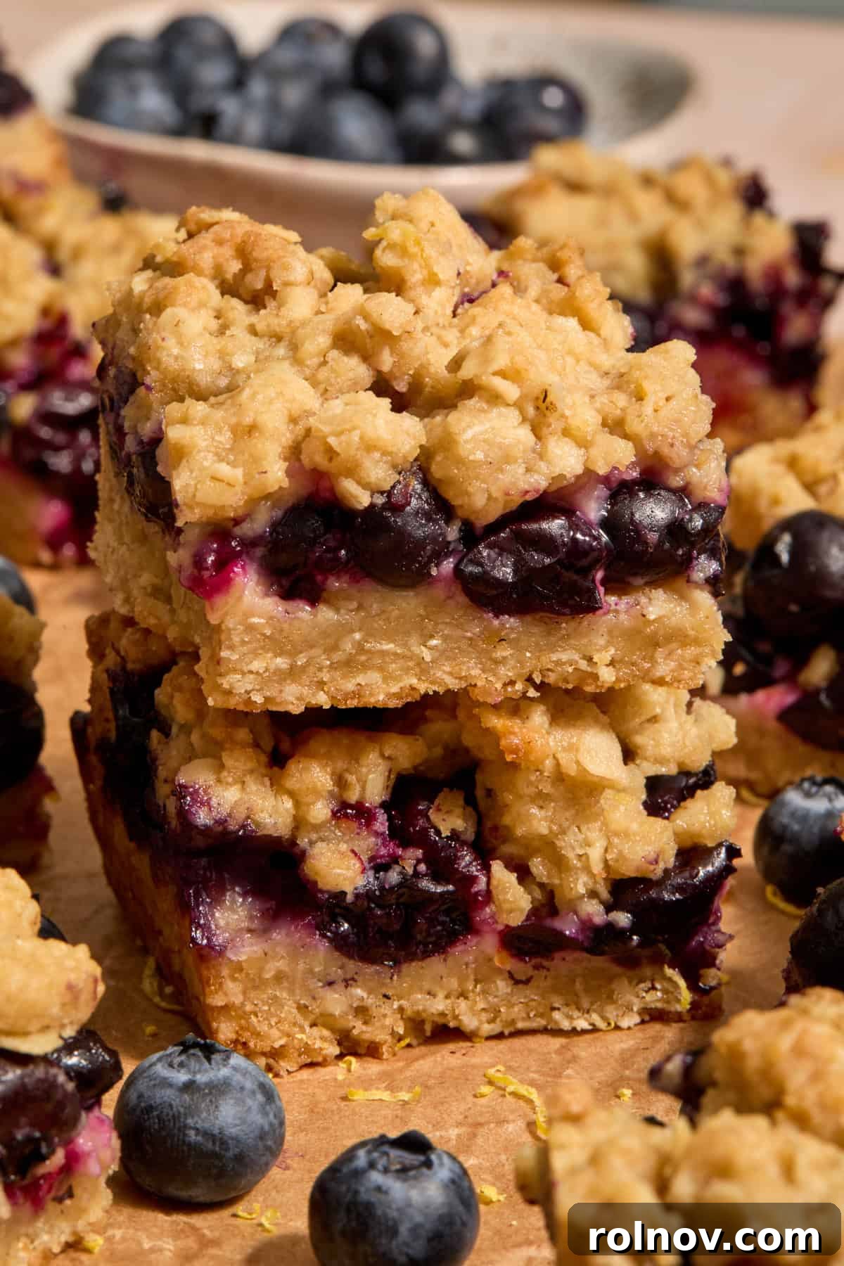 Bright Blueberry Lemon Squares 2 Stacked blueberry lemon bars on parchment paper, surrounded by fresh blueberries and lemon slices.