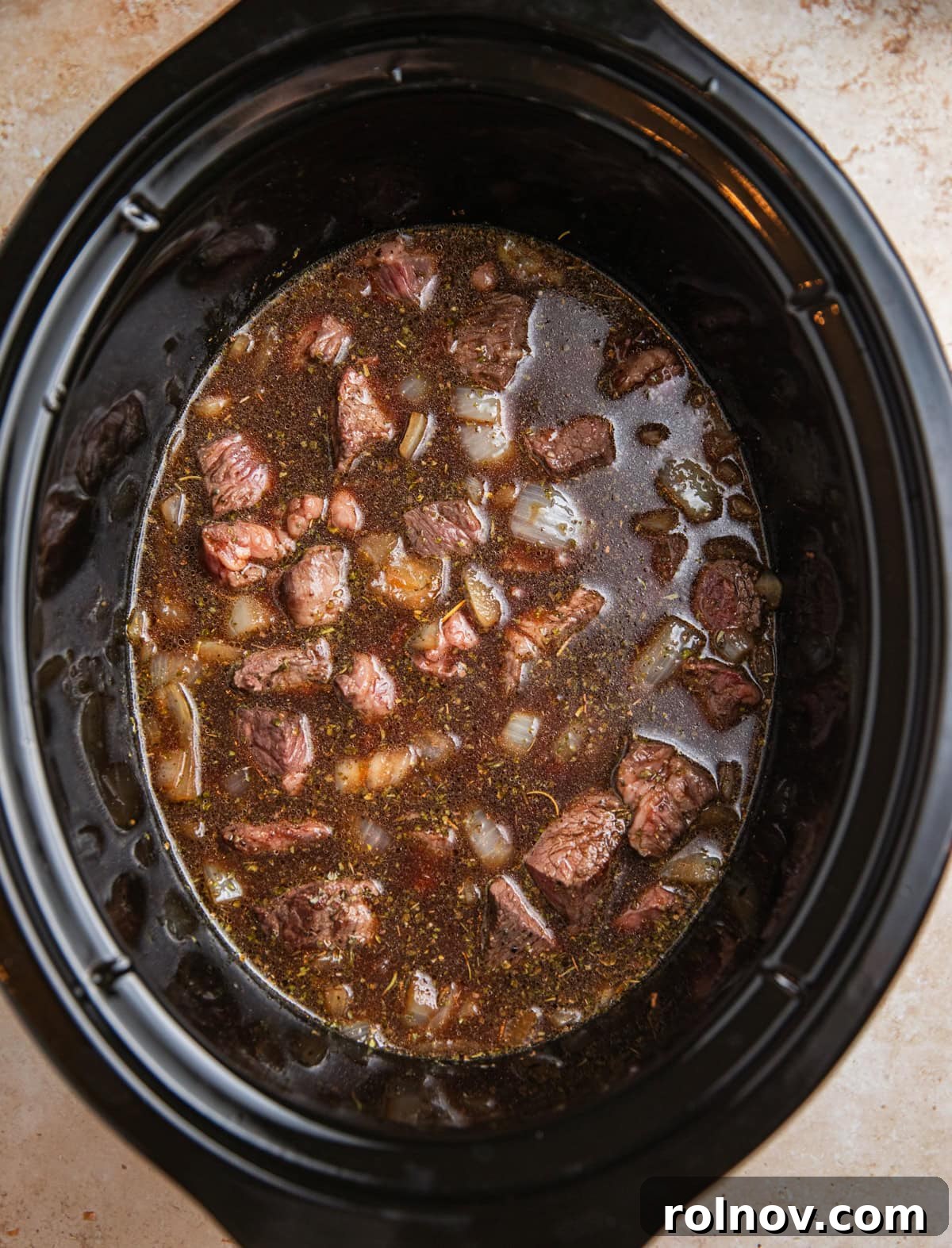 Crockpot with cubed seared beef, broth and other ingredients before slow cooking.